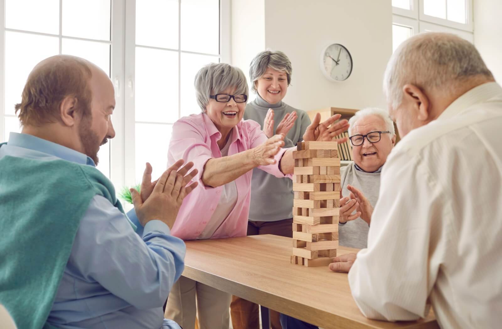 A group of senior adults, smiling and laughing, play a game of Jenga together in a bright room with large windows.